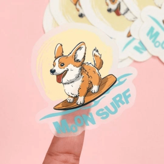 mano che tiene adesivo trasparente con stampato un cane che fa surf