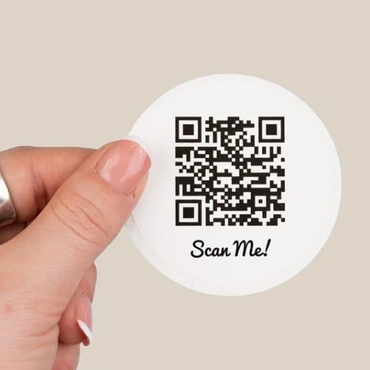 Una mano tiene un adesivo rotondo con un codice QR e la scritta 'Scan Me!'