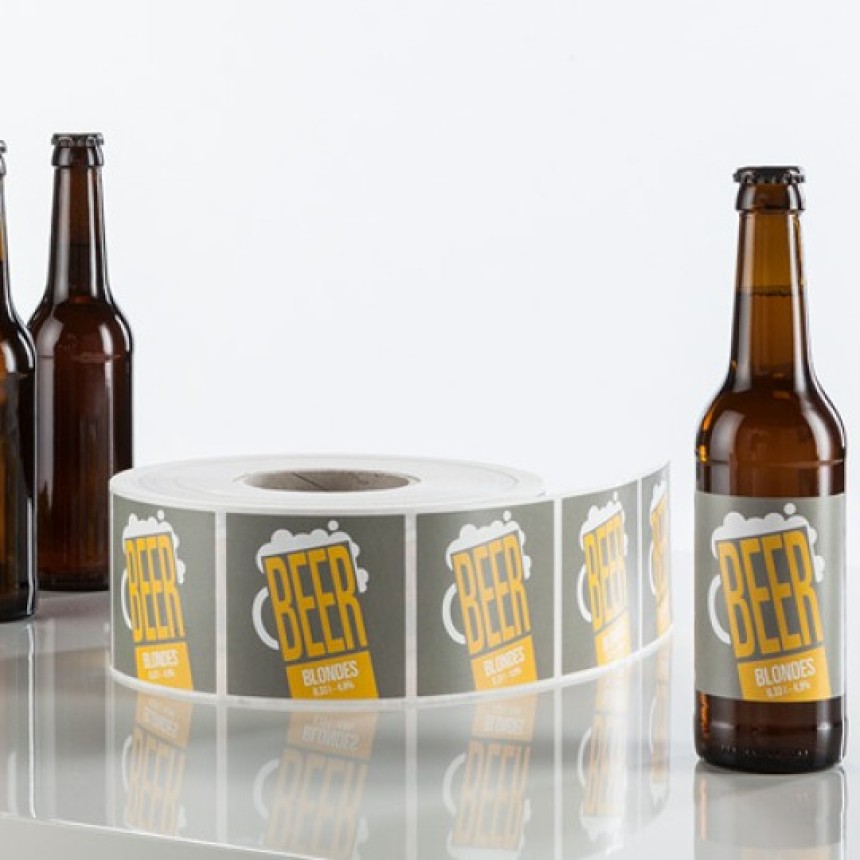 bottiglie di birra con applicate delle etichette adesive rettangolari accanto al rotolo di etichette con la stessa grafica