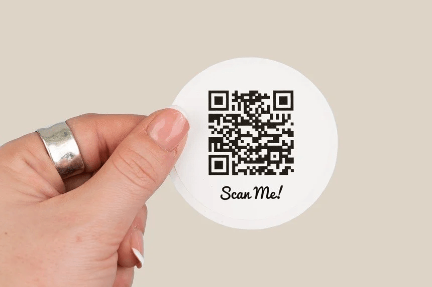Una mano tiene un adesivo rotondo con un codice QR e la scritta 'Scan Me!'