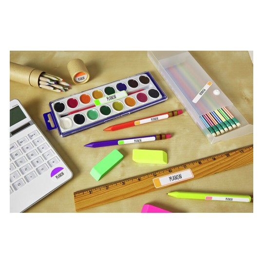 L'immagine mostra una varietà di materiali scolastici disposti su un tavolo. Ci sono acquerelli, matite colorate, evidenziatori, gomme, una calcolatrice e un righello. Tutti gli oggetti hanno etichette con il nome 'MANON'.