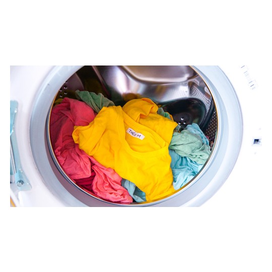 L'immagine mostra una lavatrice con abiti colorati all'interno. Ci sono magliette di vari colori, tra cui giallo, rosa e azzurro, che sembrano appena lavate.