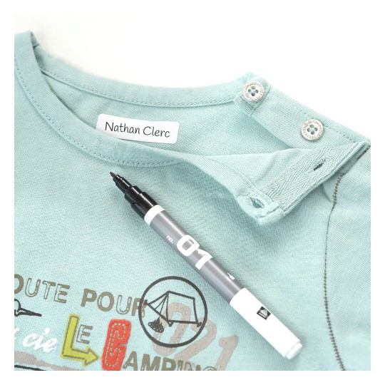 L'immagine mostra una maglietta azzurra con il nome 'Nathan Clerc' etichettato all'interno e una penna o marcatore nera posizionata accanto ad essa. La maglietta presenta anche un design grafico sul davanti, visibile in parte.