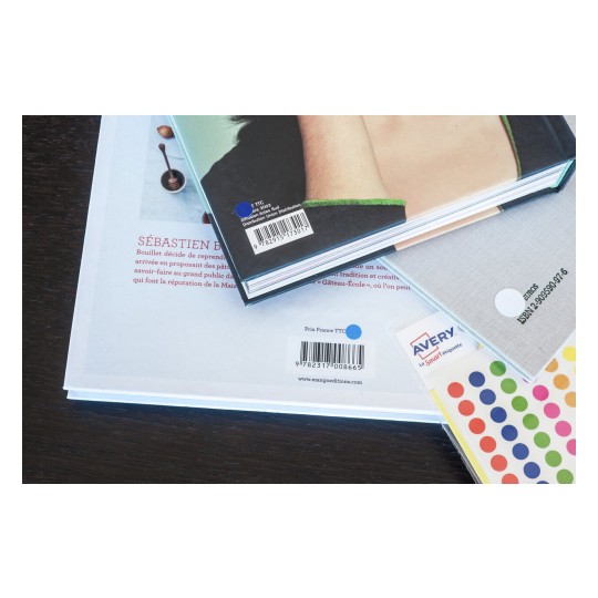 L'immagine mostra alcuni libri e un pacchetto di etichette adesive. I libri hanno copertine e codici a barre visibili, e il pacchetto delle etichette ha dei punti colorati.