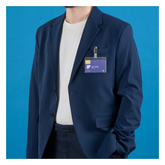L'immagine mostra una persona che indossa un abito blu scuro con una maglietta bianca. La persona ha un cartellino identificativo attaccato all'abito, e sullo sfondo c'è un colore blu uniforme.