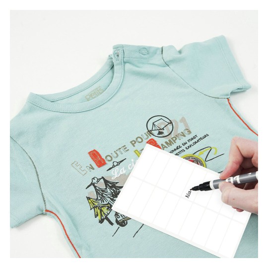 L'immagine mostra una maglietta azzurra per bambini con un design stampato. Accanto, una persona sta scrivendo su un'etichetta bianca con un pennarello.