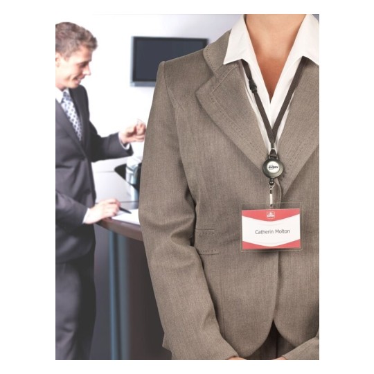 L'immagine mostra una persona in abbigliamento professionale che indossa un distintivo con un nome. Sullo sfondo, c'è un'altra persona che interagisce con un dispositivo o un computer in un ambiente d'ufficio.