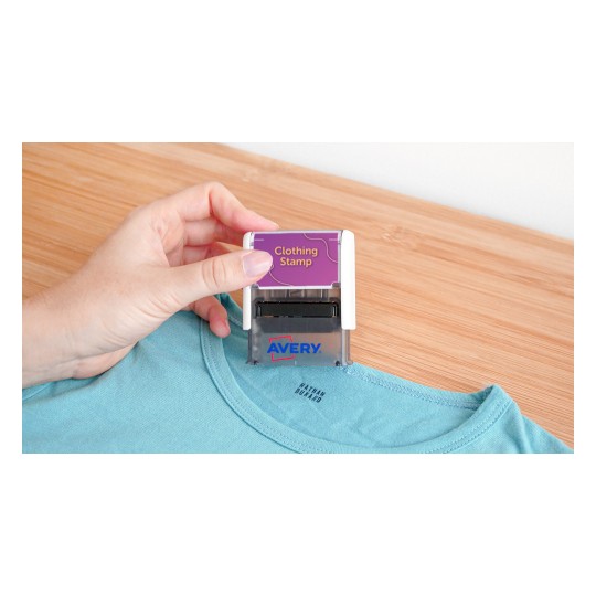 In the image, a hand is holding a clothing stamp labeled 'Clothing Stamp' from the brand Avery, positioned over the collar of a light blue t-shirt on a wooden surface.