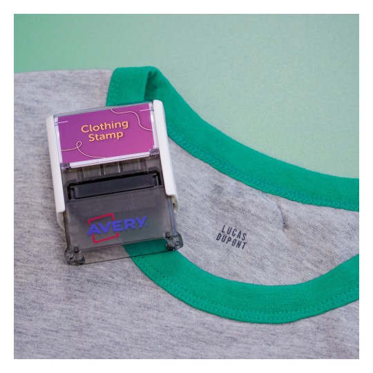 L'immagine mostra un timbro per vestiti in cima a una maglietta con colletto verde e una scritta all'interno con il nome 'LUCAS DUPONT'.