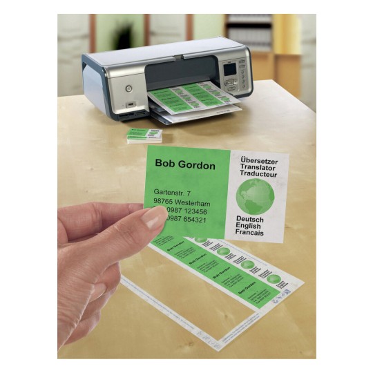 The image shows a printer on a table with printed green business cards and labels. A hand is holding one card that features contact information alongside a globe icon and multiple language translations.