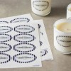 Due fogli di etichette ovali stampate con testo 'Handmade' accanto a due candele bianche su superficie grigia.
