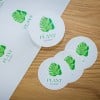 Quattro adesivi rotondi bianchi con una foglia verde e la scritta 'PLANT' su un tavolo di legno