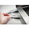 L'immagine mostra una mano che tiene un'etichetta con il nome 'ANNA', mentre un altro cavo etichettato 'KAMERA' è collegato a un laptop. I cavi sono neri e la superficie del laptop è argentata.